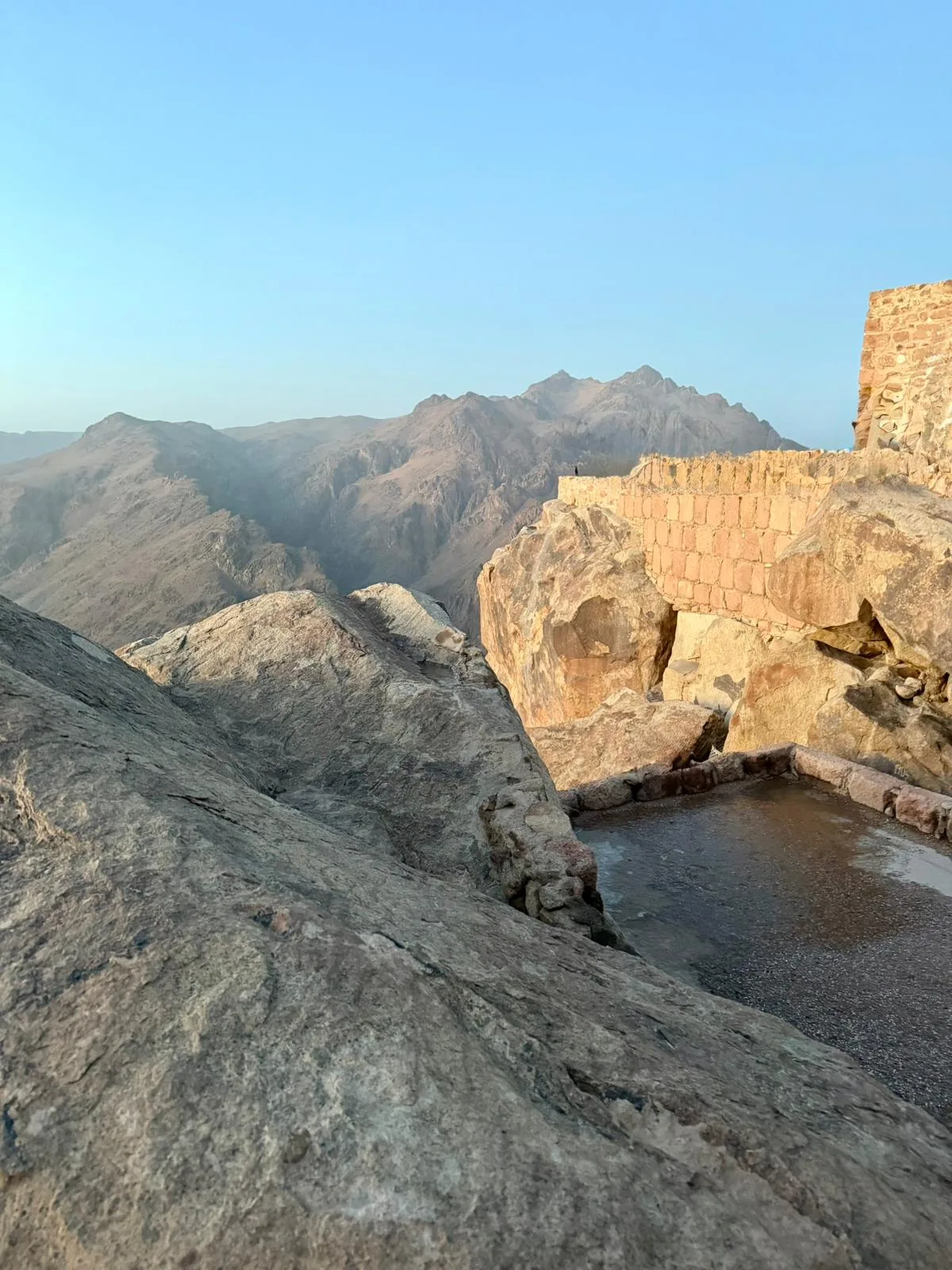 Sommet du Mont Sinaï au lever du jour, mur de pierre ancienne de la chapelle orthodoxe, blocs de granit massifs au premier plan, paysage minéral de montagnes érodées et ciel bleu clair